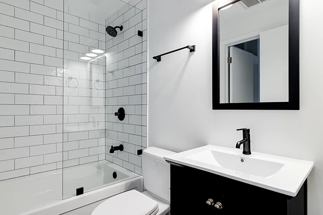 A small modern bathroom with a dark vanity, mirror frame, and hardware. White subway tiles line the bathtub and shower with black faucets