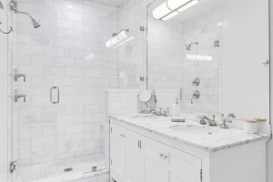 A luxurious white bathroom with a double vanity and bronze faucets, a white subway tile shower, and glass door