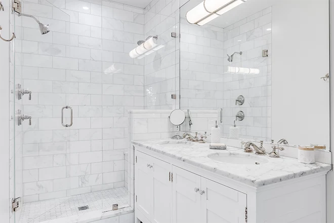 A luxurious white bathroom with a double vanity and bronze faucets, a white subway tile shower, and glass door