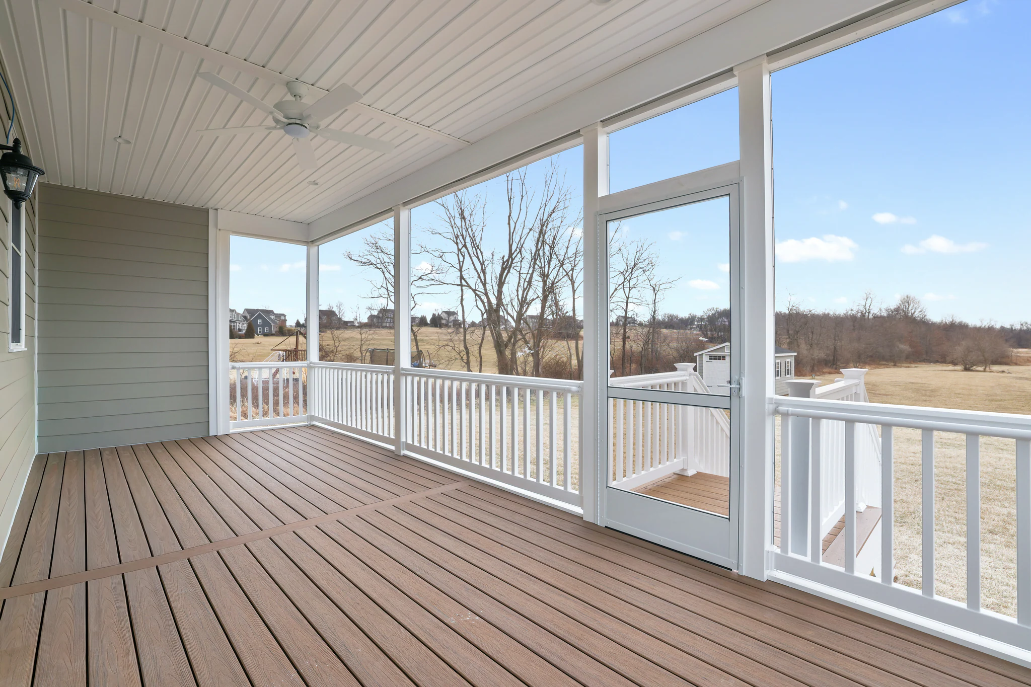Bright screened porch with white framing, wooden deck flooring, and a white ceiling fan; large windows reveal a sunny backyard with leafless trees and houses in the distance.
