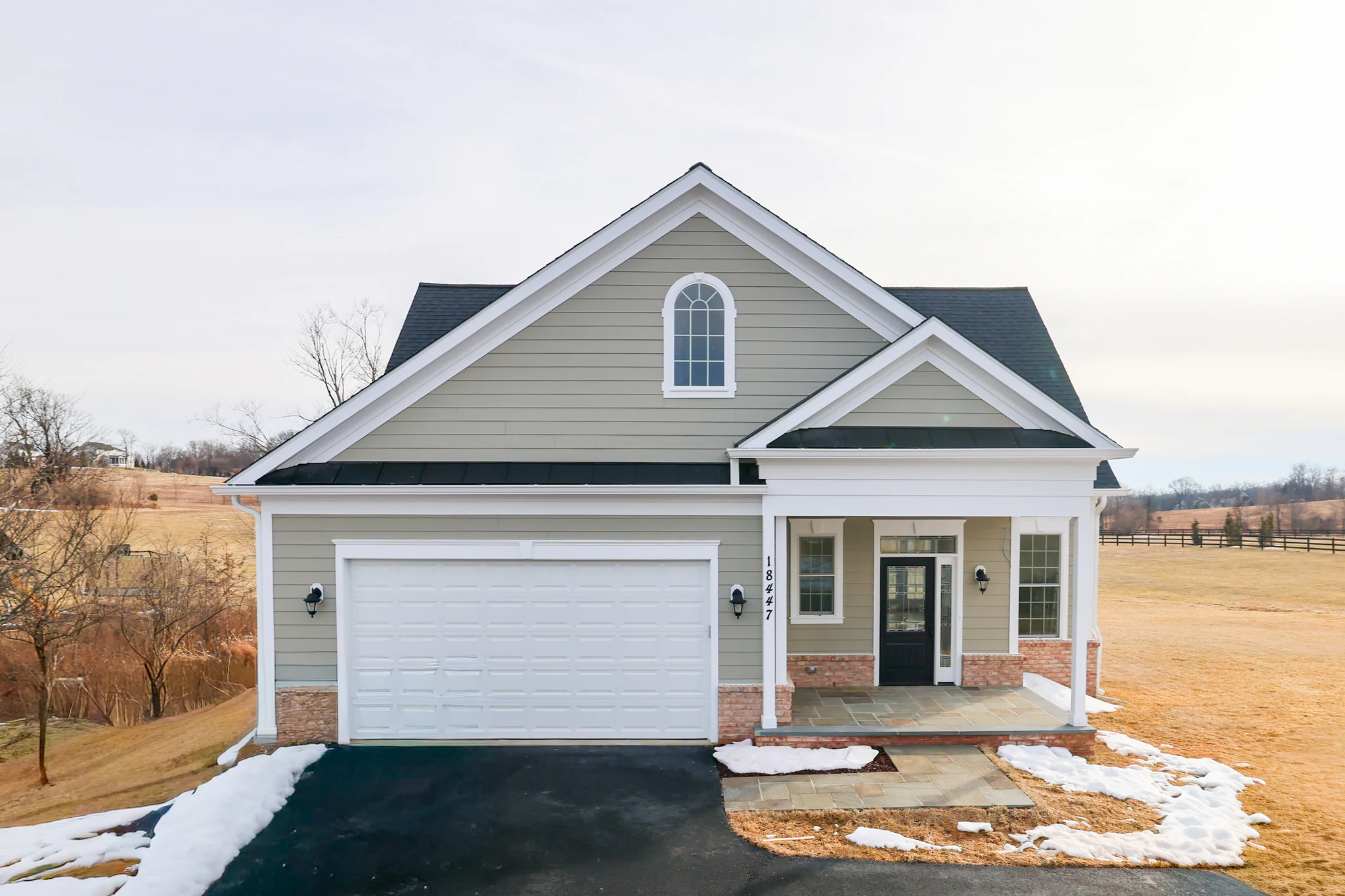 Two-story suburban house with beige siding, white trim, arched window, and attached garage on a paved driveway with patches of snow on the lawn nearby.