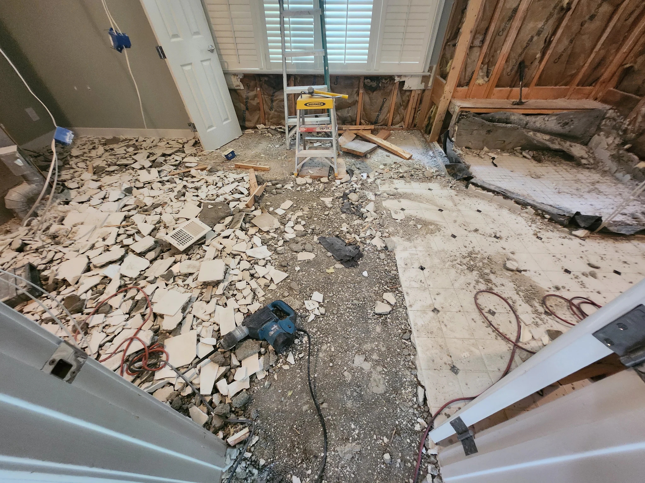 Room under renovation with exposed wall studs, damaged floor, and scattered debris including broken tiles and drywall chunks; a yellow Werner ladder sits in the center with tools nearby and coiled cords on the ground.